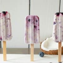 3 blueberry coconut frozen yogurt popsicles standing up with a real coconut and blueberries