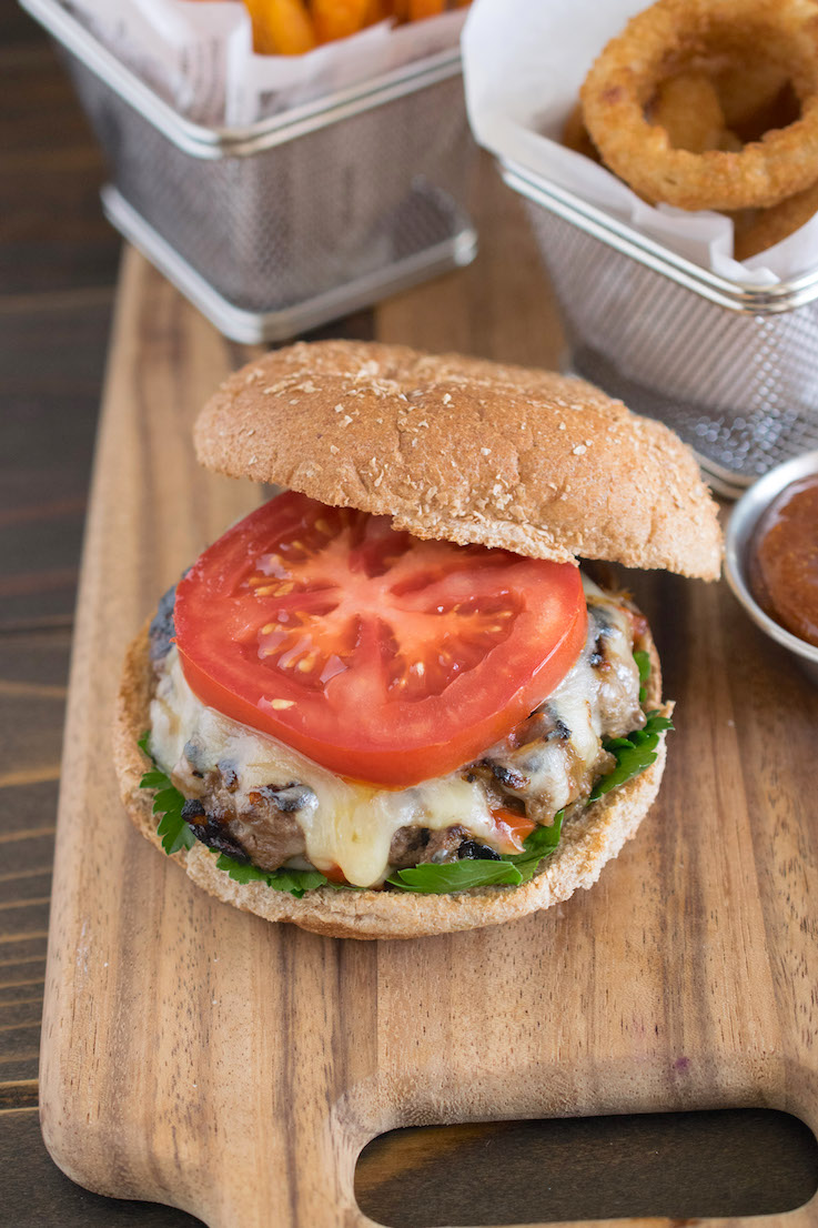 Caramelized red pepper and onion burger on a wheat bun with a slice of tomato on a board with onion rings