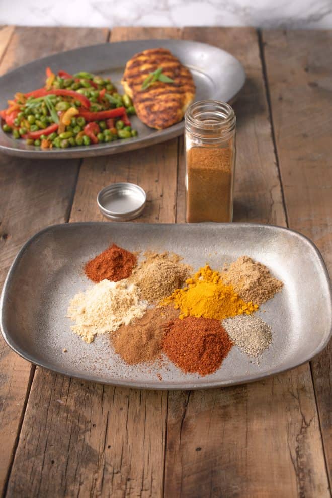 A plate with piles of different color spices with a plate of grilled chicken and peas in the background