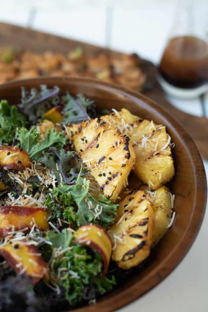 A wood bowl of green and red kale topped with grilled pineapple, shaved carrots and toasted coconut