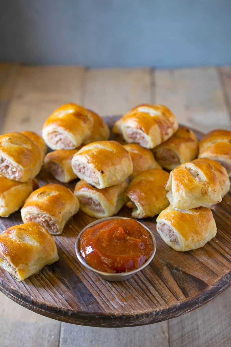 Mini pork apple & sage sausage rolls are stacked on a wooden serving board with a silver bowl of curry ketchup