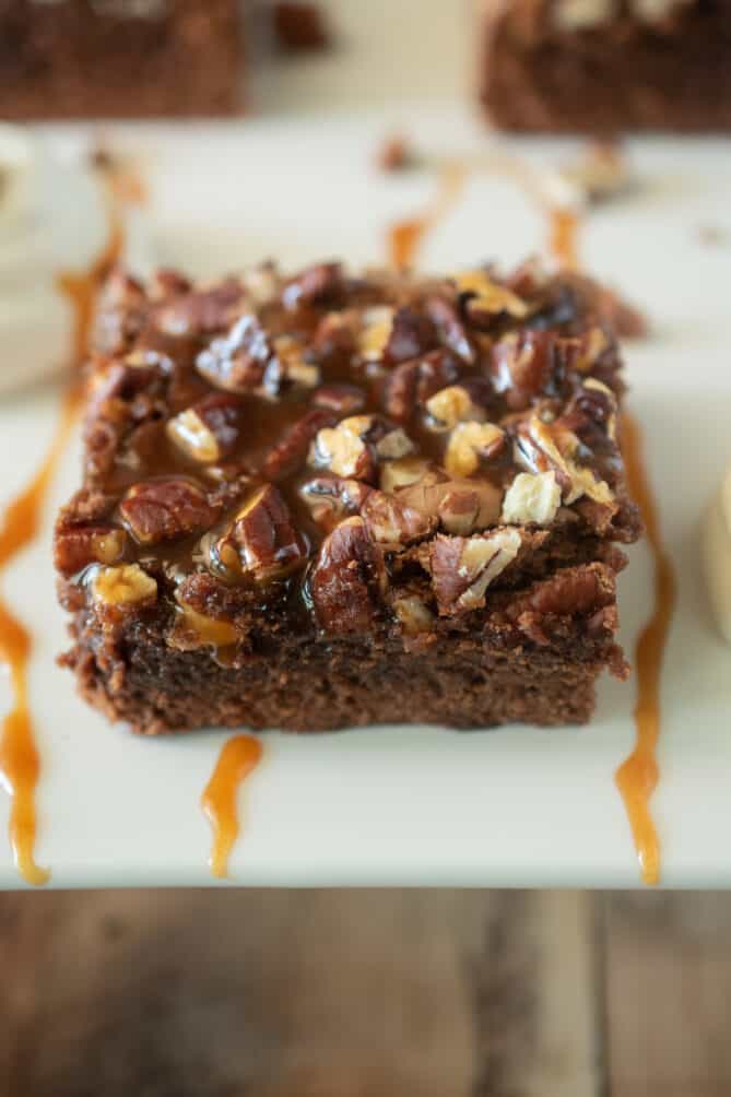 A square of salted caramel pecan brownie on a white plate with extra caramel sauce
