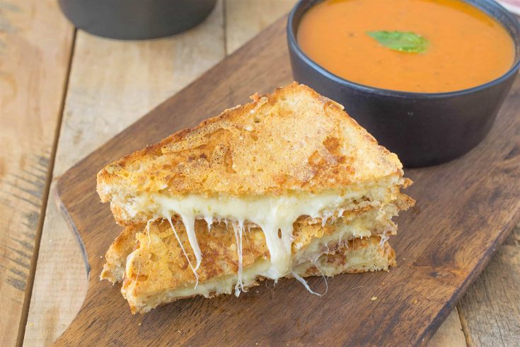 A triple grilled cheese sliced in half showing the melted cheese inside on a serving board with tomato basil soup